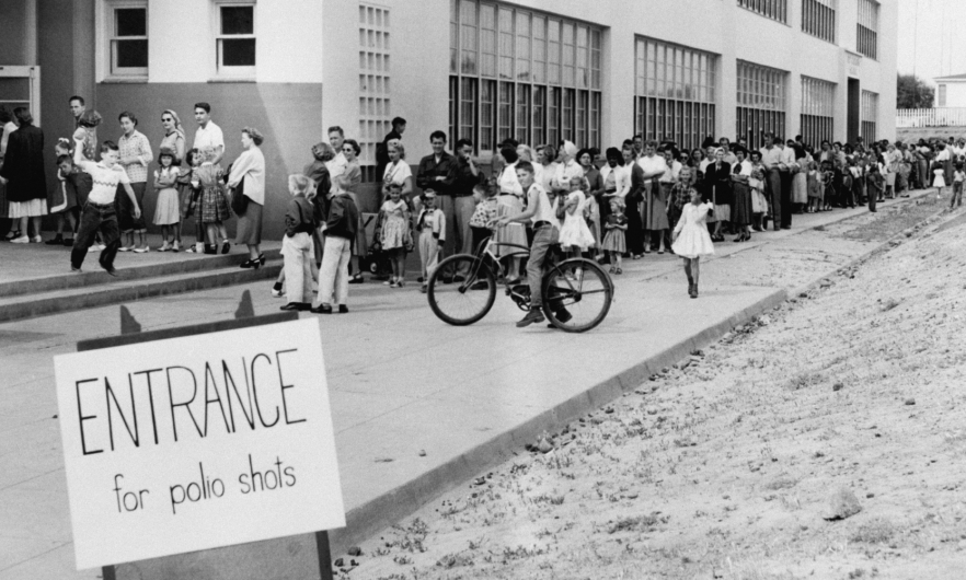 People line up for polio shots, 1955 Black and white historical photo from 1955 of families lined up at a school for polio shots. The line wraps around the building.